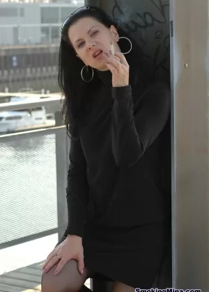 Полностью одетая брюнетка закуривает сигарету, надев на голову солнцезащитные очки